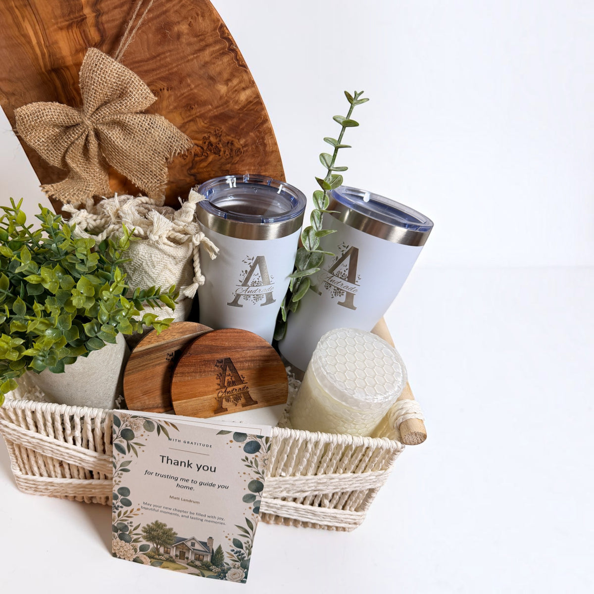 Close-up of Grand Welcome personalized closing gift basket with engraved olive wood board, tumblers and greenery